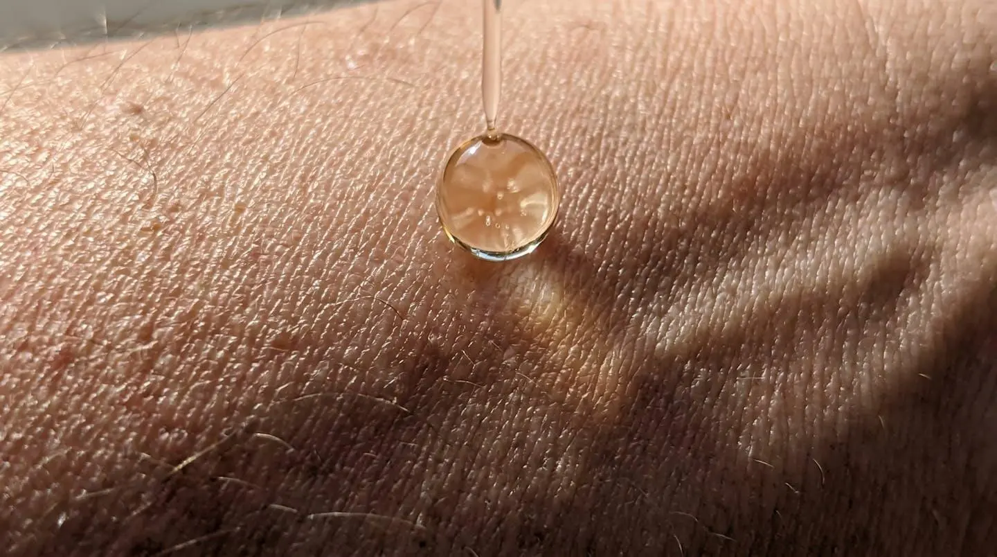 Gros plan extrême d'une goutte d'huile translucide tombant sur la peau humaine, texture cutanée naturelle visible, reflets lumineux subtils